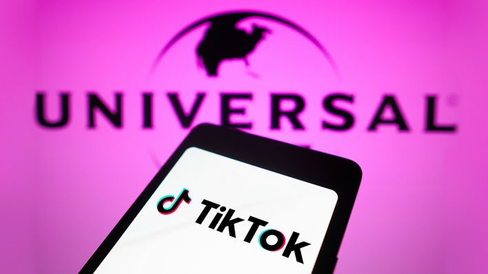 Auf einem Smartphone ist das TikTok Logo auf weißem Hintergrund zu sehen. Der Hintergrund hinter dem Smartphone ist rosa mit dem Universal Logo darauf. | Foto: IMAGO / SOPA Images