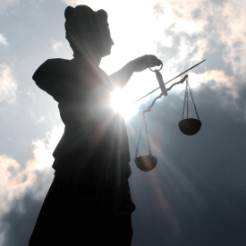 Silhouette einer Justitia-Skulptur mit Schwert und Waagschalen vor schwarzen, düsteren Wolken und Gegenlicht © imago images/Ralph Peters