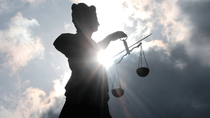 Silhouette einer Justitia-Skulptur mit Schwert und Waagschalen vor schwarzen, düsteren Wolken und Gegenlicht © imago images/Ralph Peters