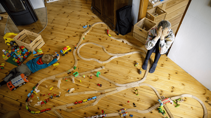 Eine gestresste Mutter sitzt verzweifelt auf dem Fußboden während ihr Kind inmitten von Spielzeug spielt © imago images/photothek