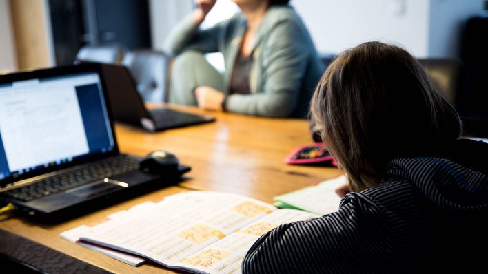 Ein Kind sitzt an einem Küchentisch und löst Aufgaben in einem Schulheft. Im Hintergrund sitzt die Mutter und arbeitet im Homeoffice.