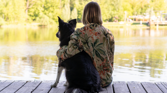 Eine Frau sitzt mit einem Hund an einem See © IMAGO/Westend61