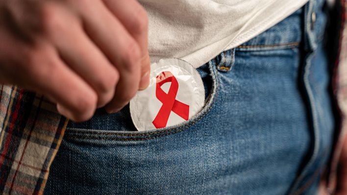 Eine Person zieht aus der Hosentasche eine Kondom-Verpackung mit der roten Aids-Schleife (Symbolbild) © dpa/Zoonar