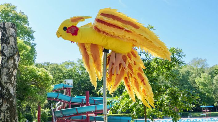 Dardan Zhegrovas große Papagei-Figur wippt hinter den Terrassen über den Köpfen der Freibadbesucher*innen © TROPEZ