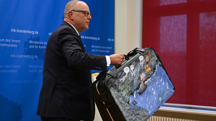 Innenminister Michael Stübgen (CDU) nimmt den Negativpreis "Abschiebeminister 2023", den er in Form eines Koffers bekommen hat, nach seinem Statement vom Tisch © dpa/Soeren Stache