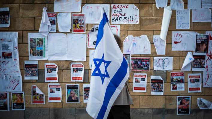Eine Frau, die eine israelische Fahne hält, guckt sich Tel Aviv Plakate an, auf denen Personen zu sehen sind, die von der Hamas entführt wurden © picture alliance/dpa | Ilia Yefimovich