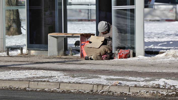 Ein obdachloser Mensch sitzt im Winter an einer Bushaltestelle in Berlin © IMAGO / Bernd Elmenthaler