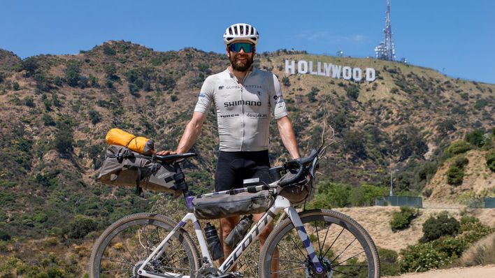 Der deutsche Athlet Jonas Deichmann steht mit seinem Fahrrad vor der Kulisse der Hollywood Hills in den USA © picture alliance/dpa/Jonas Deichmann | Markus Weinberg