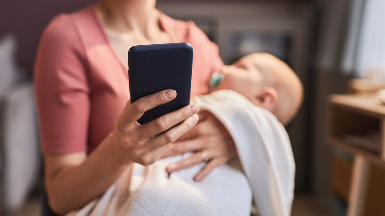 Eine Frau mit einem Baby auf dem Arm und einem Smartphone in der Hand © IMAGO / Pond5 Images