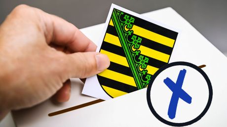 Bildmontage: Das Wappen von Sachsen wird in eine Wahlbox geworfen © IMAGO/Christian Ohde