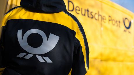 Eine Postbotin trägt vor einem Zustellfahrzeug eine Jacke mit dem Logo der Deutschen Post © imago images/Silas Stein